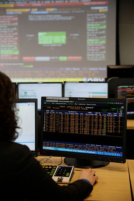 A student working at a computer with financial tables on the screen and a financial figures projected on a big screen behind the computer.