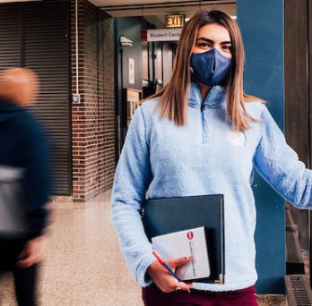 A female student in a light blue fleece jacket and dark blue mask leans her hand against the wall while people walk by the hallway behind her. 