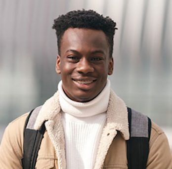 An African American male student stands outside smiling weraing a white turtleneck sweater, khaki sheep lined coat and bookbag on his shoulders. 